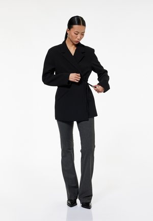 Donna che allaccia un blazer nero a portafoglio, indossa pantaloni grigi e scarpe nere con punta appuntita, in piedi su sfondo bianco.