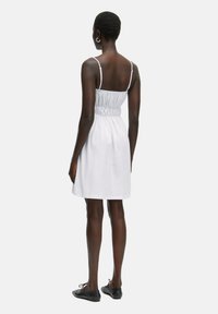 Weißes ärmelloses Kleid mit einem gerafften Oberteil und einem ausgestellten Rock, der dünne Träger und eine Länge bis zur Mitte des Oberschenkels hat. Getragen mit schwarzen flachen Schuhen.
