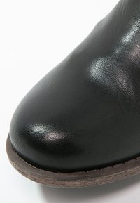 Gros plan sur la pointe d'une chaussure en cuir noir avec une texture lisse et le bord de la semelle empilée marron visible en dessous.