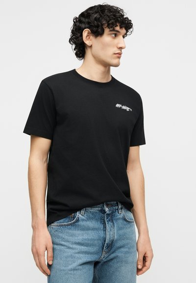 Άνδρας με σγουρά μαλλιά που φοράει κοντομάνικο μαύρο μπλουζάκι με άσπρο κείμενο στο στήθος και ανοιχτόχρωμο μπλε τζιν, κοιτάζει προς τα δεξιά του.