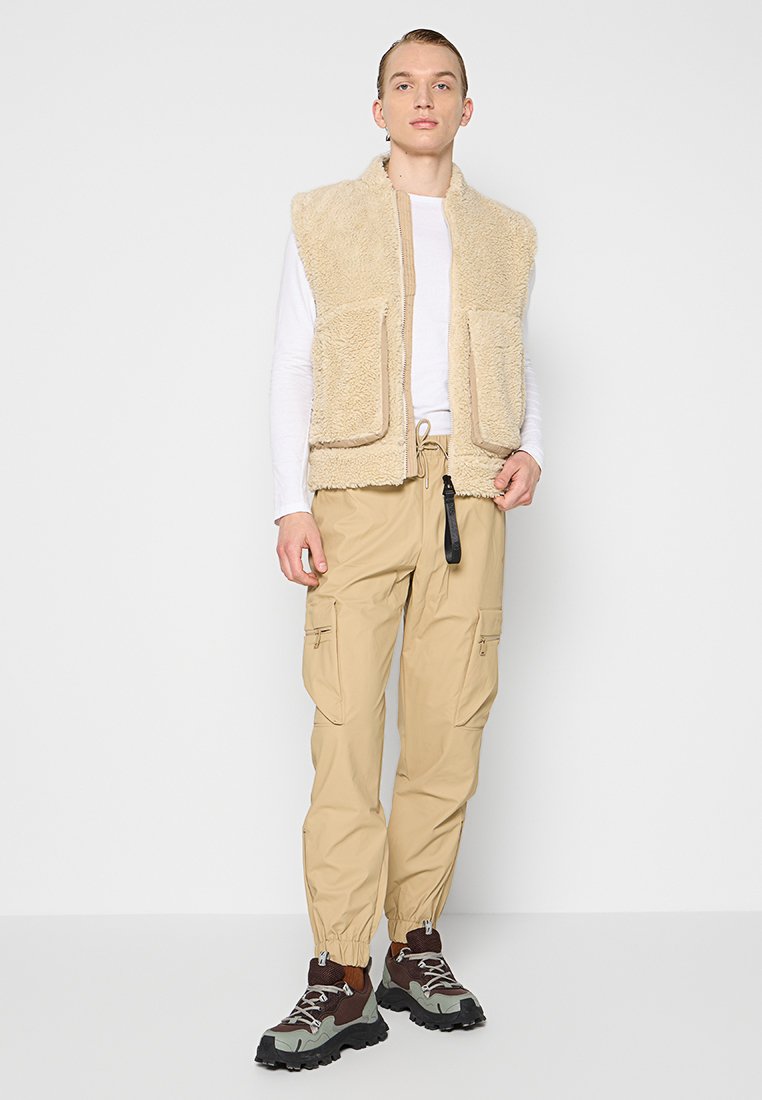 Gilet in pile beige con due tasche frontali, abbinato a pantaloni cargo di colore beige chiaro con polsini elastici e vita con cordoncino. Stivali da escursionismo scuri.