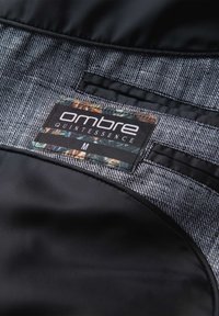 Étiquette de vêtement portant la mention "ombre QUINTESSENCE M", cousue à l'intérieur d'un tissu gris et noir avec détail floral sur l'étiquette.