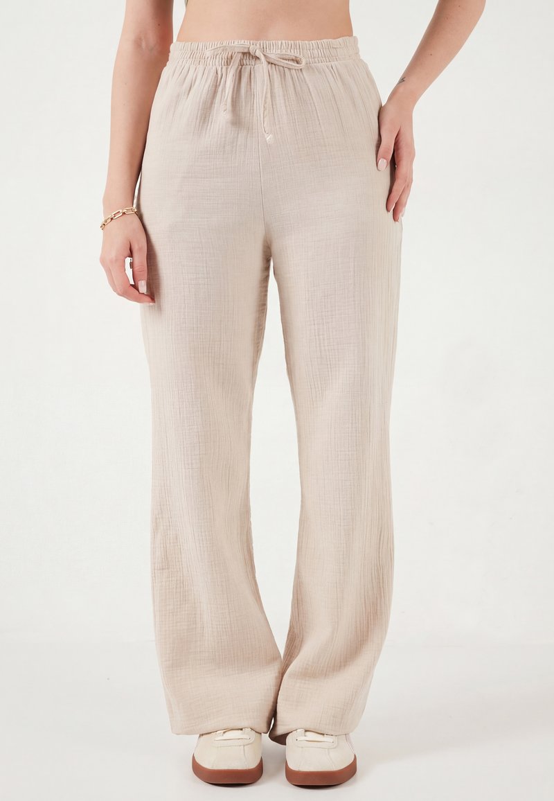 Naine, kes kannab beežikat nööriga sääretaga tekstuurse kangaga pükse ja kreemjaid tossusid, seisab tavalise valge tausta ees.