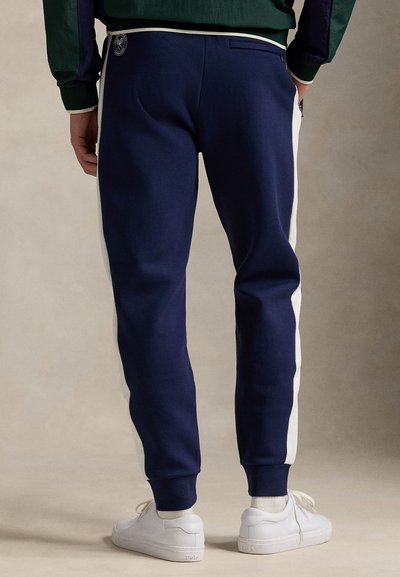 Marinblå joggingbyxor med vita sidor och ribbade muddar. Mjukt tyg och en bakficka förstärker designen. Bärs med vita skor.