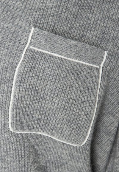 Šedá pletená tkanina zobrazuje detailní pohled na náprsní kapsu s bílým lemováním. Textura se jeví jako ribbovaná s měkkou, chlupatou kvalitou.