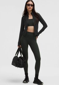 Crna zip-up sportska jakna, zeleni sportski grudnjak i usklađene tajice. Model drži crnu torbu za sport, nosi elegantne sunčane naočale i patike.