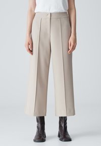 Pantalons beige à large jambe avec plis avant, taille haute et fermeture à bouton unique, associés à des bottines brunes.