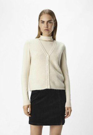 Ljust beige ribbat cardigan med V-hals, knappstängning och lång ärm, lager på lager över en ljusbeige polotröja, kompletterad med en svart manchesterkjol.