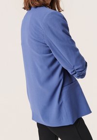 Modrý blejzr z texturované látky, s přiléhavým střihem, kleslými ramenními švy a jednou postranní kapsou.