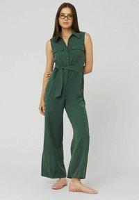 Combinaison verte sans manches avec un col, des boutons, des poches latérales et une taille nouée. Portée avec des ballerines ouvertes rose. Texture de tissu lisse.