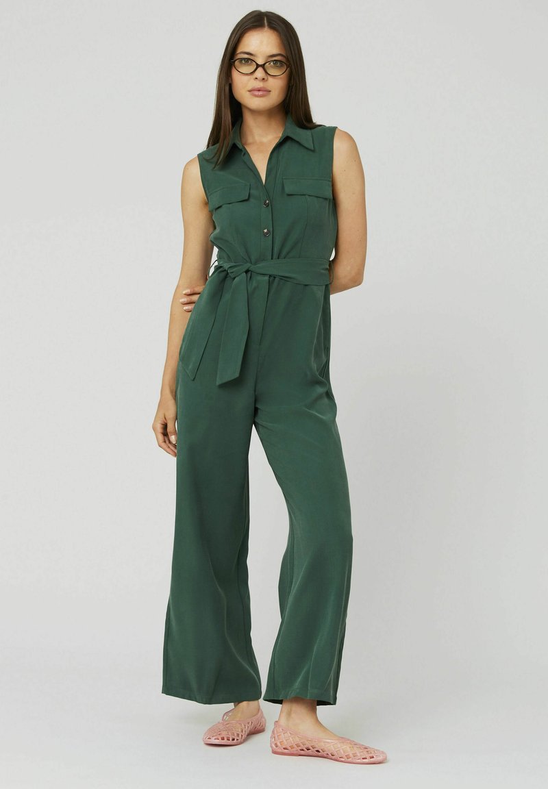 Combinaison verte sans manches avec un col, des boutons, des poches latérales et une taille nouée. Portée avec des ballerines ouvertes rose. Texture de tissu lisse.