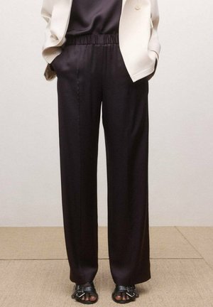 Personne portant un pantalon noir ample à taille élastique, un blazer beige et des sandales noires à bout ouvert avec des détails à boucle, debout sur un sol beige.