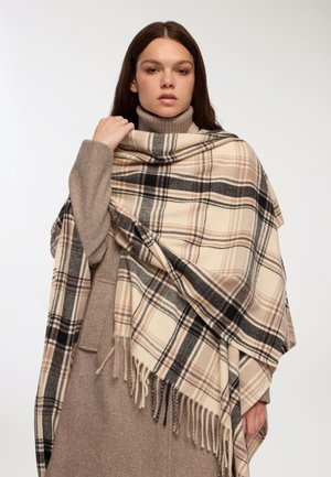 Sciarpa in misto lana a quadri nei toni beige, marrone e nero con bordi frangiati; abbinata a un dolcevita marrone chiaro e un capo esterno.