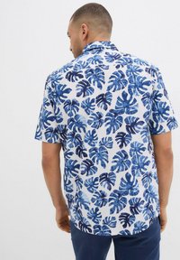 Camicia a maniche corte in tessuto bianco con stampa di foglie tropicali blu. Vestibilità rilassata con colletto classico e dettaglio sulla parte posteriore.