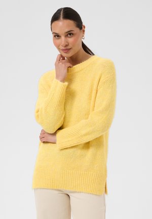 Femme aux cheveux foncés attachés en queue de cheval porte un pull jaune large et un pantalon clair, debout devant un fond clair uni, la main près du menton.