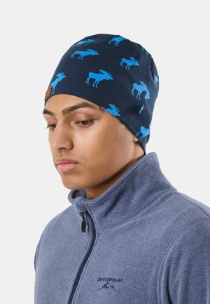 Jeune homme portant un bonnet bleu marine avec des imprimés d'élans bleus et une veste en polaire à fermeture éclair bleu clair.