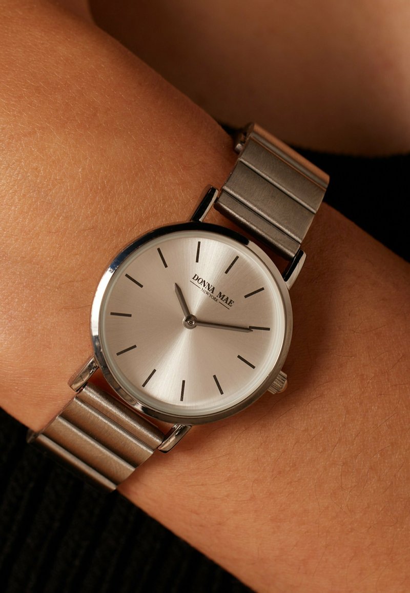 Armbanduhr mit silbernem Metallarmband und minimalistischem silbernem Zifferblatt, das 10:14 zeigt, getragen an einem Handgelenk mit brauner Haut und schwarzem Ärmel.