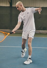 White tennis shirt with red accents, white shorts, and black and white athletic shoes. Player swings a tennis racket on a blue court.
