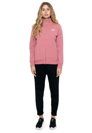 Une femme se tient devant un fond blanc, vêtue d’un sweat-shirt rose zippé, d’un pantalon noir et de baskets noires.