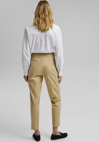 Pantaloni beige sartoriali con vestibilità slim, dotati di tasche posteriori. Abbinati a una camicia bianca con bottoni, mostrati di spalle.