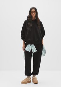 Haut à capuche noir oversize avec une chemise rayée verte et blanche nouée à la taille, assorti à un pantalon noir et des chaussures plateformes beiges avec des lanières.