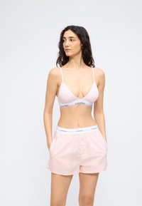 Γυναίκα με σκούρα σγουρά μαλλιά που φοράει ανοιχτό ροζ bralette Calvin Klein και σορτς που ταιριάζουν, όρθια με τα χέρια στις τσέπες, κοιτάζοντας αριστερά.