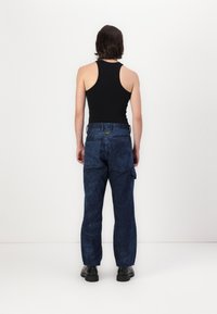 Persona de espaldas, vistiendo una blusa sin mangas negra, pantalones azules con estampado y grandes bolsillos, y botas negras gruesas, sobre un fondo blanco.