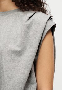 Grå kortermet topp laget av mykt stoff; har en avslappet passform med subtile skulderfolder og en båthals.