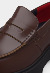 Καφέ δερμάτινο παπούτσι τύπου penny loafer με ραφή μοκασίνου και κόκκινη εσωτερική επένδυση σε μαύρη σόλα, φαίνεται σε κοντινό πλάνο.