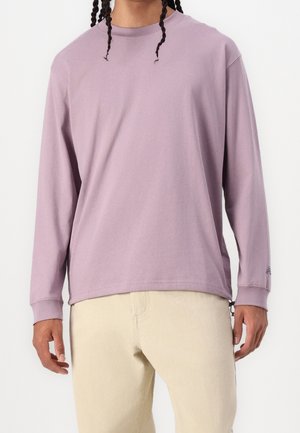 Mężczyzna z warkoczami, ubrany w jasnofioletową koszulę z długim rękawem i jasnobeżowe spodnie, stojący na tle jednolitego tła.