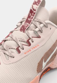 Lyse beige Nike Trail sneakers med tekstureret mesh-overdel, maroon accenter, mønstrede snørebånd og signaturlogo på siden.