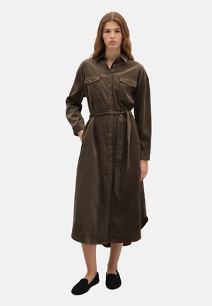 Donna che indossa un vestito midi marrone a maniche lunghe con bottoni, tasche sul petto e cintura in vita, abbinato a scarpe slip-on nere.