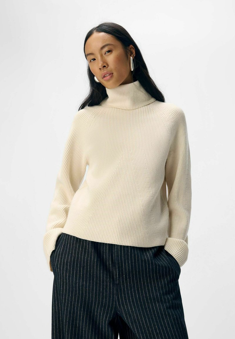 Krem turtleneck pulover z jazzbešimi rokavi, ki se ponaša z teksturiranim vzorcem. V kombinaciji s temnimi širokimi hlačami z navpičnimi črtami.