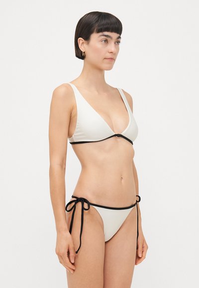 Baltais bikini komplekts ar trīsstūra topu un sānu saišu biksēm, akcentēts ar melnu apdari. Gluda auduma tekstūra, minimālistisks dizains.