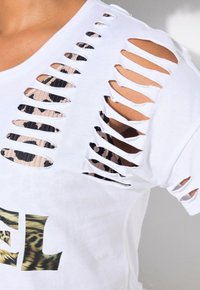 Valkoinen T-paita, jossa on revitty olkadesign, joka paljastaa alla olevan kuvioidun kankaan. Edessä rohkea monivärinen graafinen kuvio.