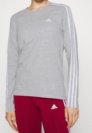 Osoba nosząca szarą koszulkę Adidas z długim rękawem z białymi paskami na rękawach oraz czerwone spodnie Adidas z białymi paskami.