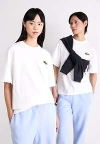 Tricouri din bumbac alb cu logo-ul brodat al crocodilului verde. Modelul poartă pantaloni scurți albaștri; al doilea model are un hanorac închis aruncat pe umeri.