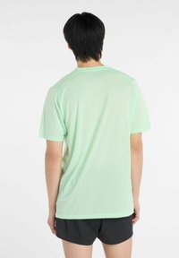 Persona de espaldas con una camiseta de manga corta verde claro y pantalones cortos negros frente a un fondo blanco liso.