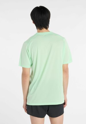 Person, der mit dem Rücken zur Kamera steht, trägt ein hellgrünes T-Shirt mit kurzen Ärmeln und schwarze Shorts vor einem schlichten weißen Hintergrund.