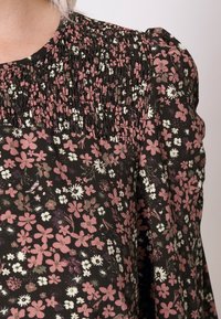 Blouse fleurie à fond noir, ornée de fleurs roses, blanches et violettes. Fronces texturées au niveau du col et manches bouffantes.