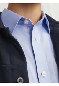 Oblečená modrá košile s límečkem pod tmavou bundou, s bílými knoflíky a hladkým povrchem. Viditelné švy a detaily kovových prvků na bundě.