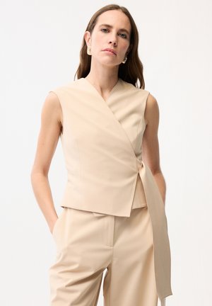 Donna con capelli castani lunghi che indossa un top avvolgente beige senza maniche e pantaloni abbinati, mani in tasca, sfondo neutro.