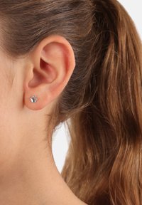 Orecchino a forma di corona argentata con tre accenti sferici. Design piccolo, adatto per un look casual, indossato all'orecchio di un modello.