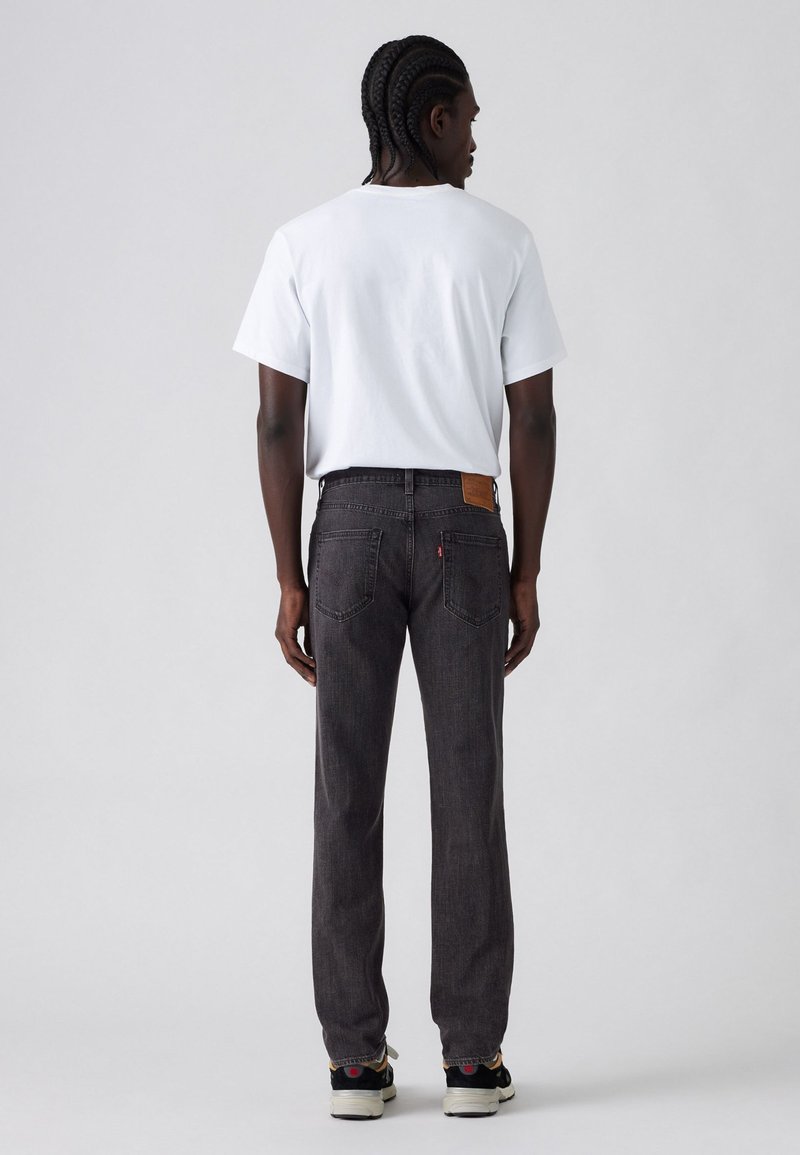 Jeans de mezclilla negra con corte recto, tejido texturizado y una parche de cuero marrón en la cintura trasera, combinados con una camiseta blanca.