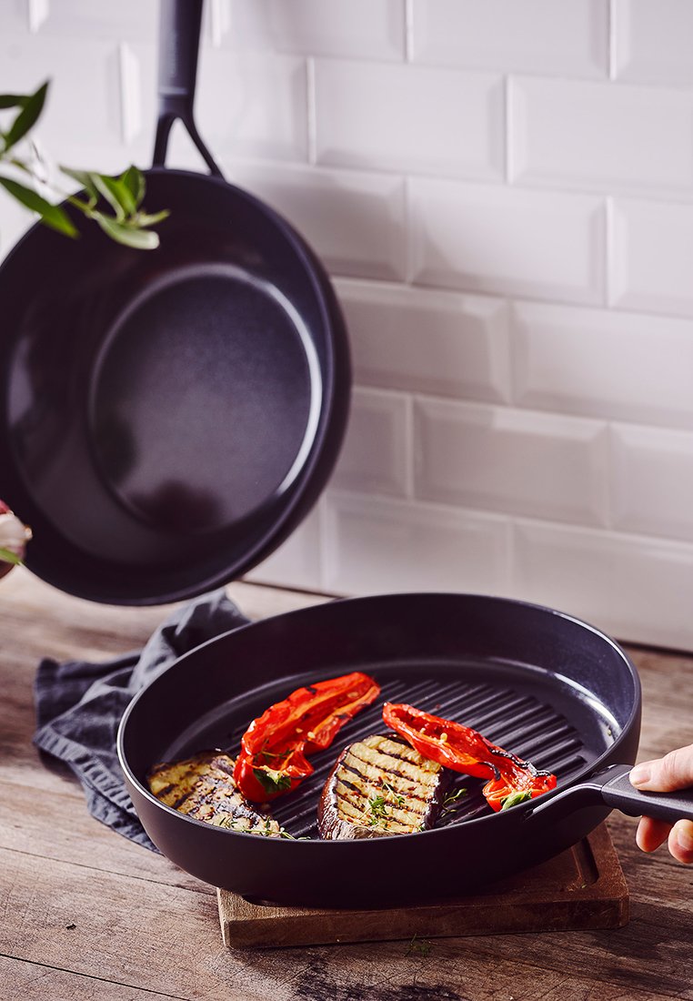 Sartén de parrilla negra antiadherente con una superficie lisa y acanalada, con verduras a la parrilla y pimientos rojos, sobre una tabla de cortar de madera.