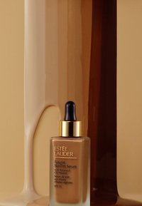 Flacon compte-gouttes en verre brun avec des accents dorés, contenant un sérum teinté. Texture lisse, SPF 20, sur un fond dégradé beige.