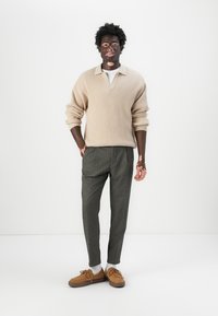 Maglione beige a costine, pantaloni grigi su misura, mocassini marroni con lacci e calze bianche. Il modello è in piedi contro uno sfondo bianco.