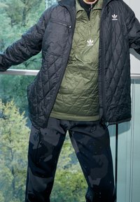 Svart quiltad jacka över en grön pufftröja, kombinerad med svarta camouflagebyxor med färgglada inslag. Alla plagg har synliga varumärkeslogotyper.