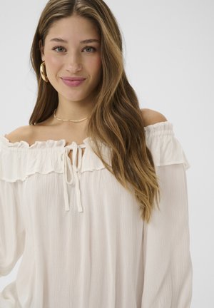 Krémově bílá halenka s odhalenými rameny, zdobená volánkovým lemem a zavazováním u výstřihu. Měkká, texturovaná látka s volnými, splývavými rukávy.