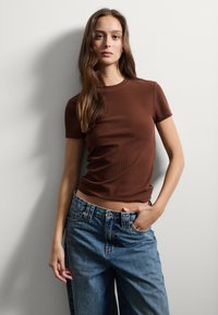 Bruine geribbelde top met korte mouwen, gecombineerd met losse blauwe spijkerbroeken. De stof lijkt textuur te hebben met een aansluitende silhoutte bij de taille.